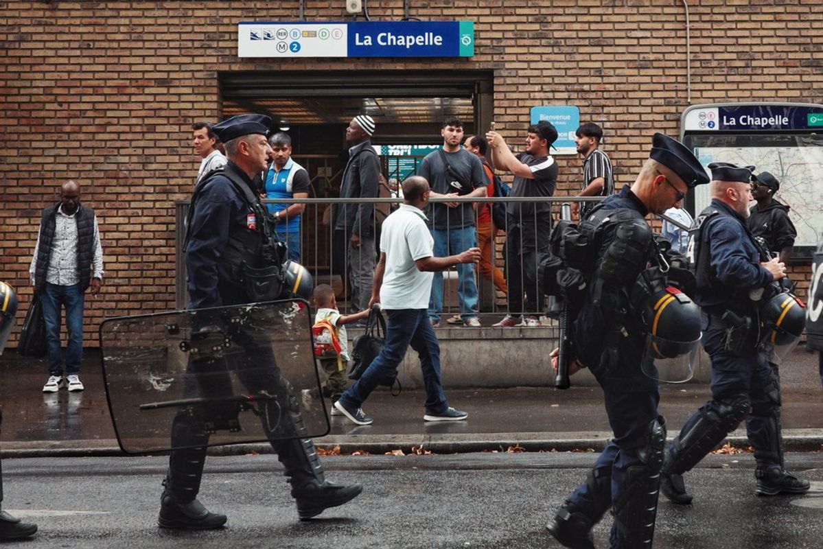 Riot police walking down the street in front of the La Chappelle métro station on Paris. People at the entrance are looking to the left and taking photos.